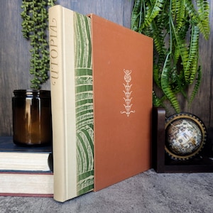 May include: A vintage hardcover book titled "Utopia" with a green and white patterned spine and a solid brown cover. The book is sitting on a table with other books and a glass jar.