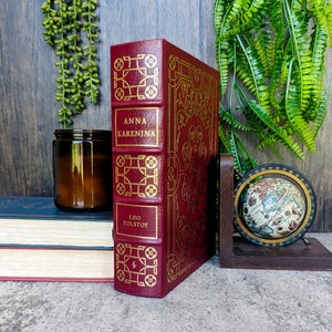 May include: A burgundy hardback book titled "Anna Karenina" by Leo Tolstoy, with gold embossed lettering and decorative patterns. The book is propped up by a wooden bookend, next to a small globe and a candle.