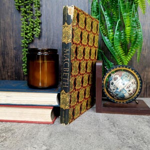 May include: A copy of Shakespeare's "Macbeth" with a gold and red patterned cover stands between a wooden bookend. A brown candle in a glass jar and a small globe are also visible. The scene is set against a backdrop of greenery and other books.