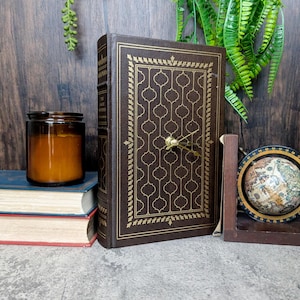 Puede incluir: Un reloj decorativo con forma de libro marrón con un patrón geométrico dorado. Las manecillas del reloj son doradas. También se ven un pequeño globo terráqueo y una vela en un tarro de cristal.