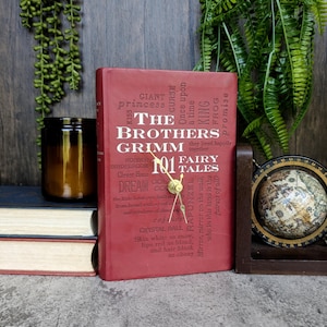 May include: A red book with a gold clock face on the cover. The book is titled "The Brothers Grimm 101 Fairy Tales". The cover is decorated with words related to fairy tales, such as "giant", "princess", "curse", "once upon a time", "king", "frog", and "promise".
