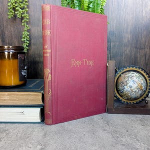 May include: A maroon hardcover book titled "Ebb-Tide" by Christian Reid. The book has gold lettering on the spine and cover. A stack of books and a candle are visible in the background.