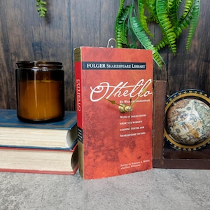 Könnte beinhalten: Ein rotes Buch der Folger Shakespeare Library mit "Othello" von William Shakespeare und einem Uhrwerk. Eine braune Glas-Kerze und ein kleiner Globus sind ebenfalls zu sehen. Das Buch hat detaillierte Notizen.