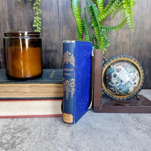 Puede incluir: Una pila de libros antiguos, con un libro azul en el centro con letras doradas y una pequeña etiqueta. También se ven un pequeño globo terráqueo y una vela marrón en un tarro. El fondo incluye vegetación y una superficie de madera.