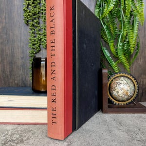 Pode incluir: Um livro encadernado em vermelho com o título "THE RED AND THE BLACK" impresso na lombada. O livro está sobre uma mesa com uma superfície de madeira.