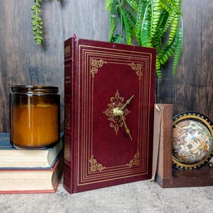Pode incluir: Um relógio em forma de livro na cor bordô com detalhes dourados em filigrana. O relógio tem um mostrador e ponteiros dourados. Outros itens incluem uma vela em um pote, um pequeno globo e uma pilha de livros.