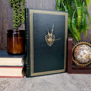 May include: A green book with gold accents and a clock face on the front. The clock face has gold hands and a gold shield with a design in the center. The book is sitting on a table with other books and a globe.