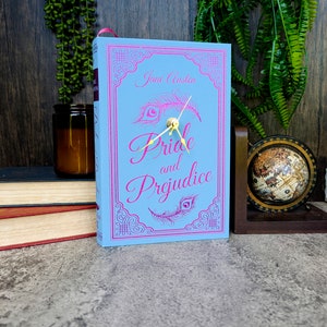 May include: A blue book clock with a gold clock face and hands. The book cover features the title "Pride and Prejudice" by Jane Austen in pink lettering with a peacock feather design.