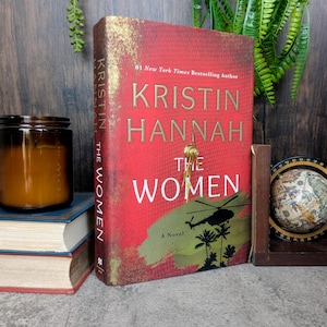 May include: A hardcover book titled "THE WOMEN" by Kristin Hannah. The cover is red with gold lettering and a silhouette of a helicopter and palm trees. A candle and a stack of books are in the background.