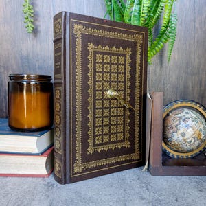 May include: A brown, antique book with gold embossed detailing and a decorative clasp. The book is standing upright, next to a candle in a glass jar, a small globe, and other books. The book's cover features a repeating square pattern.