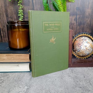May include: A green hardcover book titled "THE WINE-PRESS" by Alfred Noyes. The book has gold lettering and a gold illustration of a bird. Other books, a candle, and a globe are in the background.