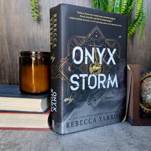 May include: A hardcover book titled "Onyx Storm" by Rebecca Yarros, with a black cover and gold accents. The book is propped up against other books and a candle, with green foliage in the background.