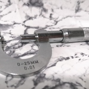 以下が含まれることがあります： 0〜25mm、精度0.01mmのシルバーメタル製のノギス。ノギスは白い大理石の表面に置かれています。