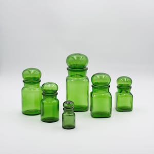 Pode incluir: Uma coleção de seis frascos de boticário de vidro verde com tampas arredondadas. Os frascos, de tamanhos variados, apresentam um design consistente: um corpo cilíndrico ou retangular e uma tampa a condizer. Dispostos sobre uma superfície branca.