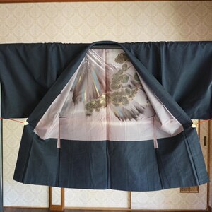 May include: A dark navy kimono is displayed on a black metal rack. The interior lining is a light pink with a detailed gold and grey floral design. Red tassels hang from the sleeves, and the kimono is open, showcasing the intricate pattern.