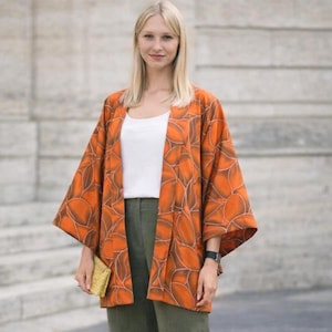 Könnte beinhalten: Eine orangefarbene Jacke im Kimono-Stil mit braunem Blättermuster wird über einem weißen Oberteil und olivgrünen Hosen getragen. Eine kleine goldene Clutch und weiße Ballerinas vervollständigen das Outfit. Die Ärmel der Jacke sind weit.
