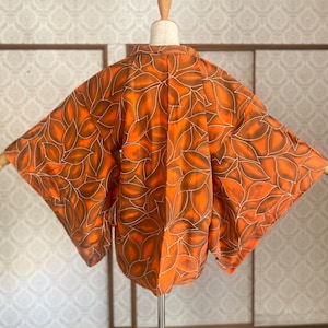 Peut inclure: Kimono orange avec un motif de feuilles marron et des contours blancs. Le vêtement est présenté sur un mannequin, mettant en valeur ses larges manches et son design fluide. L'arrière-plan présente un mur à motifs et un écran japonais traditionnel.