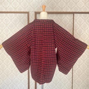 May include: A red and black patterned kimono-style jacket displayed on a mannequin. The jacket features a repeating circular design in red against a black background. The sleeves are wide and the collar is visible.