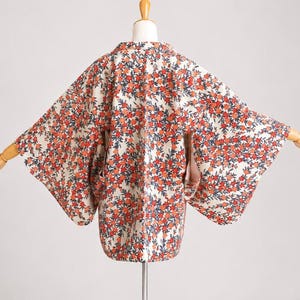May include: A floral kimono in cream with a pattern of orange and navy flowers. The kimono is displayed on a mannequin, showing its wide sleeves and open front design. The fabric appears to be lightweight and suitable for layering.