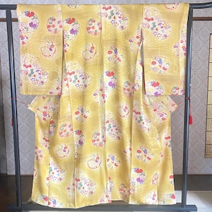 May include: A yellow kimono with a floral pattern. The kimono is displayed on a black metal rack. The floral pattern includes red, purple, and white flowers. The background is a light yellow color.