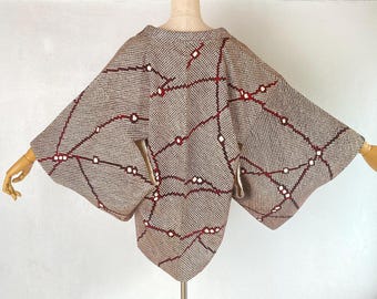 May include: A beige and brown textured jacket featuring a grid-like pattern of dark red lines and small white squares. The garment has wide, loose sleeves and a structured collar, displayed on a mannequin to show its shape and woven fabric detail.
