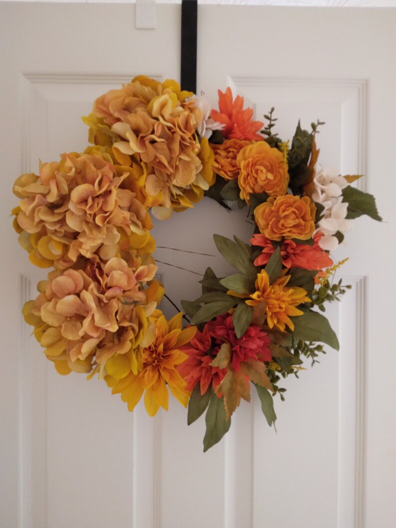 Pu&ograve; includere: Una ghirlanda autunnale con fiori arancioni, gialli e rossi. La ghirlanda &egrave; realizzata con fiori e verde artificiali.