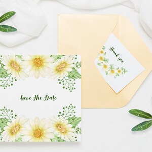 May include: A white card with a floral border and the text "Save The Date" in green script. A smaller white card with a floral border and the text "Thank you" is on top of a gold envelope.