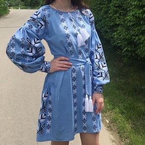 Puede incluir: Un vestido azul claro con mangas largas y abullonadas y bordados blancos y negros intrincados. El vestido presenta una cintura anudada y detalles de borlas blancas. El diseño incluye patrones geométricos y florales.