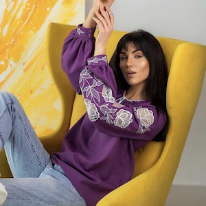 Puede incluir: Una blusa morada de manga larga con bordados florales blancos en las mangas. Se combina con vaqueros azul claro. La persona est&aacute; sentada en un sill&oacute;n amarillo, con una pintura abstracta amarilla y blanca de fondo.