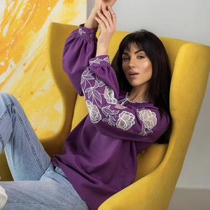 Puede incluir: Una blusa morada de manga larga con bordados florales blancos en las mangas. La blusa se combina con vaqueros azul claro. La persona est&aacute; sentada en un sill&oacute;n amarillo, con una pintura abstracta amarilla y blanca de fondo.