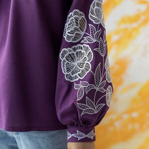Puede incluir: Una blusa morada oscura con bordados florales blancos en las mangas. Las mangas son largas y fruncidas en la mu&ntilde;eca. El dise&ntilde;o floral incluye flores grandes y hojas. La blusa se combina con unos vaqueros azules.