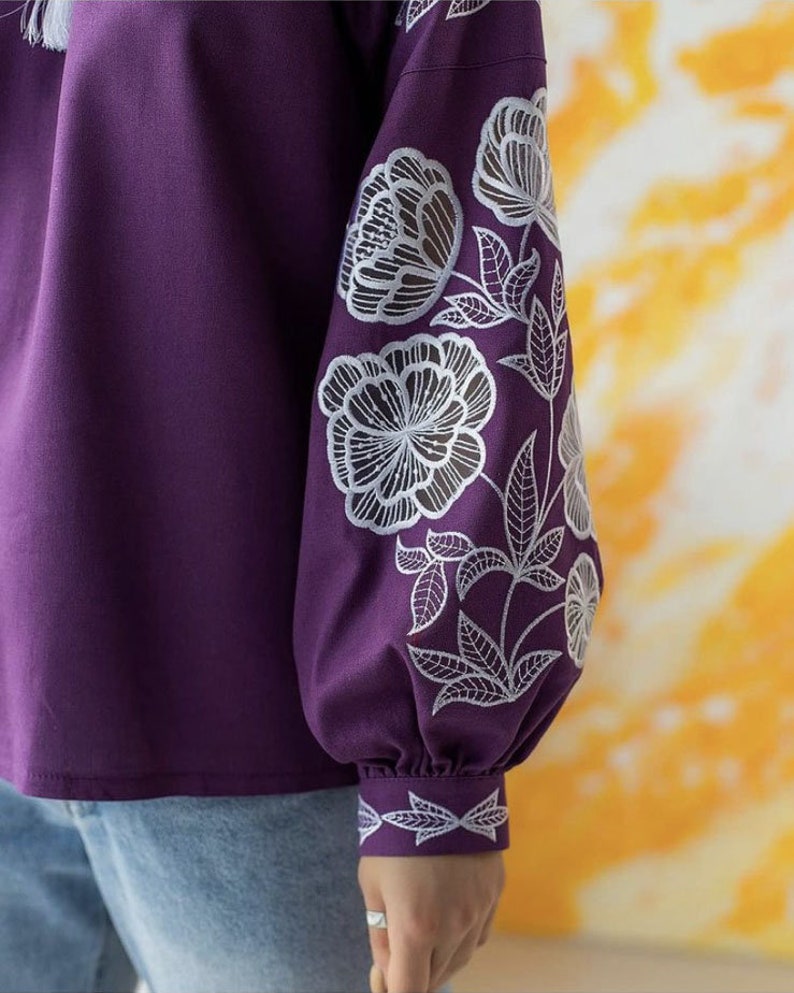Puede incluir: Una blusa morada oscura con bordados florales blancos en las mangas. Las mangas son largas y fruncidas en la mu&ntilde;eca. El dise&ntilde;o floral incluye flores grandes y hojas. La blusa se combina con unos vaqueros azules.