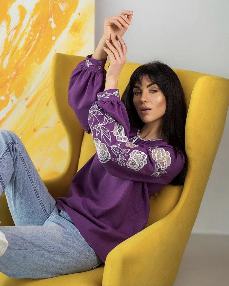 Puede incluir: Una blusa morada de manga larga con bordados florales blancos en las mangas. La blusa tiene un cuello redondo. La persona lleva vaqueros azul claro y est&aacute; sentada en un sill&oacute;n amarillo.