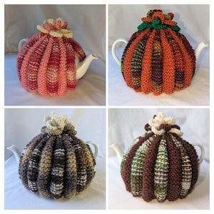 May include: Four white ceramic teapots each covered by a ribbed, hand-knitted cozy. The cozies feature varied color palettes including pink, orange, brown, and green, with each topped by a small, crocheted flower-like decorative accent.