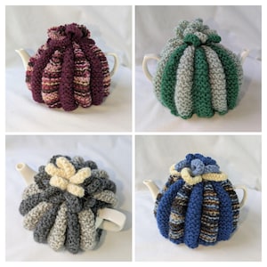 Könnte beinhalten: Vier weiße Teekannen aus Keramik, die jeweils mit einer handgestrickten Teekannenwärmer-Hülle bedeckt sind. Die Hüllen weisen vertikale Rippenstrukturen in verschiedenen Farbkombinationen auf, darunter Violett, Grün, Grau und Blau.