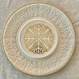May include: A white ceramic plate with a gold rim and intricate gold design. The plate has a raised center with a smaller gold design.