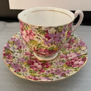 May include: A floral teacup and saucer set with a pink and purple floral pattern on a white background. The cup and saucer have a gold trim.