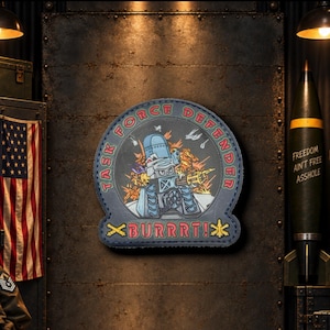May include: A circular embroidered patch featuring a blue tank character firing a weapon amidst orange explosions. The text reads TASK FORCE DEFENDER in a circular border and BURRRT! at the bottom, flanked by two yellow crossed-cannon icons.