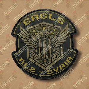 May include: A black and gold embroidered patch featuring a double-headed eagle design. The eagle is holding three stylised bullets in its talons. The text "Eagle Ruz Syria" is written around the eagle.