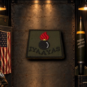 May include: A square olive green embroidered patch featuring a black bomb icon with a red flame at the top. The text IYAAYAS is stitched in black thread across the bottom of the patch, which has a dark border and a textured fabric surface.