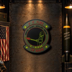 May include: A circular olive green wall sign featuring a black center with a yellow outline of a robot firing a weapon. The text reads IF IT FLIES IT DIES, COUNTER-ROCKET-ARTILLERY-MORTAR, and C-RAM, accented with four yellow stars.