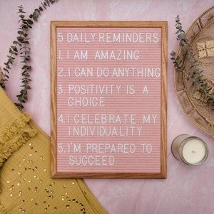 May include: A pink letter board with a wooden frame displays five daily reminders: "I am amazing", "I can do anything", "Positivity is a choice", "I celebrate my individuality", and "I'm prepared to succeed".