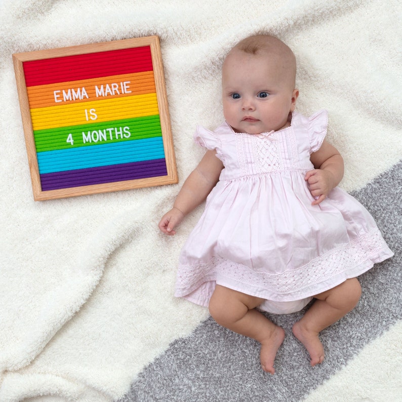 May include: A baby girl wearing a pink dress with a white lace trim. A rainbow letter board says "Emma Marie is 4 months".
