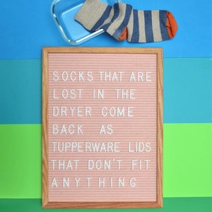 May include: A pink felt letter board with a white wooden frame. The board has the following message: "Socks that are lost in the dryer come back as Tupperware lids that don't fit anything."