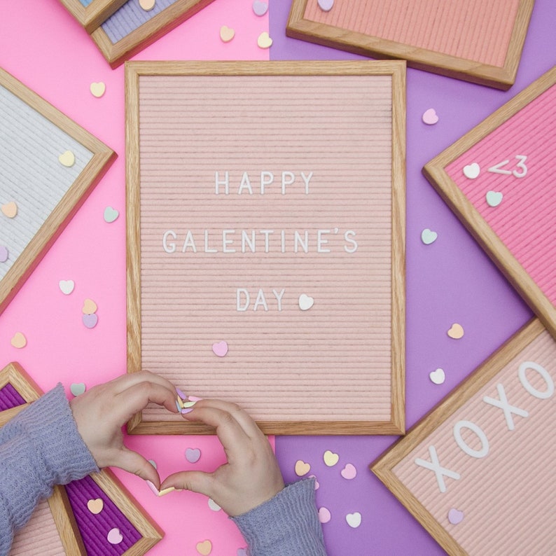 May include: A pink letter board with the text "HAPPY GALENTINE'S DAY" in white letters. The board is surrounded by pink and purple surfaces with scattered heart-shaped candies.