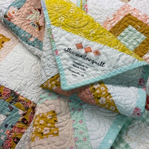 May include: A white quilt with a yellow, pink, and blue floral pattern. The quilt is folded in half and has a label that reads "attic window quilt Handmade by Sarah Fulks Huntsville, AL 11-2021".