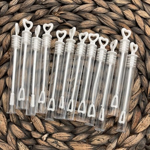 May include: A set of 12 clear plastic bubble wands with white heart-shaped handles. The wands are arranged in a row on a woven brown surface.