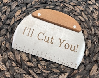 Peut inclure: Une spatule à pâtisserie en acier inoxydable avec une poignée en bois. Elle a une règle sur le fond avec des mesures en centimètres. La spatule porte la mention "I'll Cut You!" gravée.