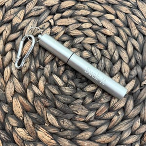 May include: A silver-colored, cylindrical metal straw with a carabiner clip and a key ring. The text "Suck It" is engraved on the side. The straw is lying on a woven, brown surface.