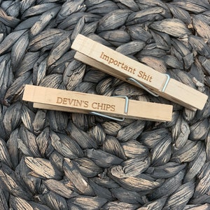 Puede incluir: Dos pinzas de madera con el texto "Important Shit" y "Devin's Chips" grabado en ellas. Las pinzas están sobre una superficie tejida marrón y negra.